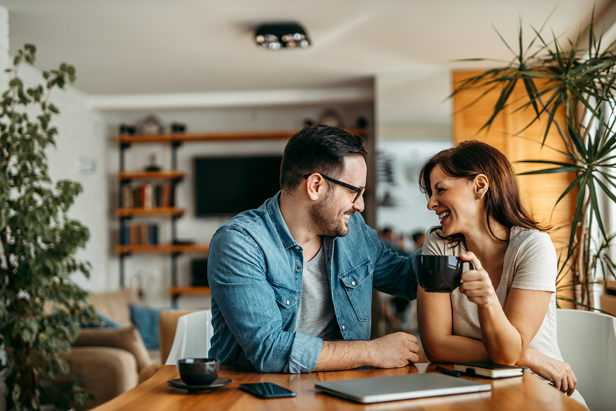 Couple enjoying coffee, sharing a laugh. They are sitting in sunny room surrounded by plants