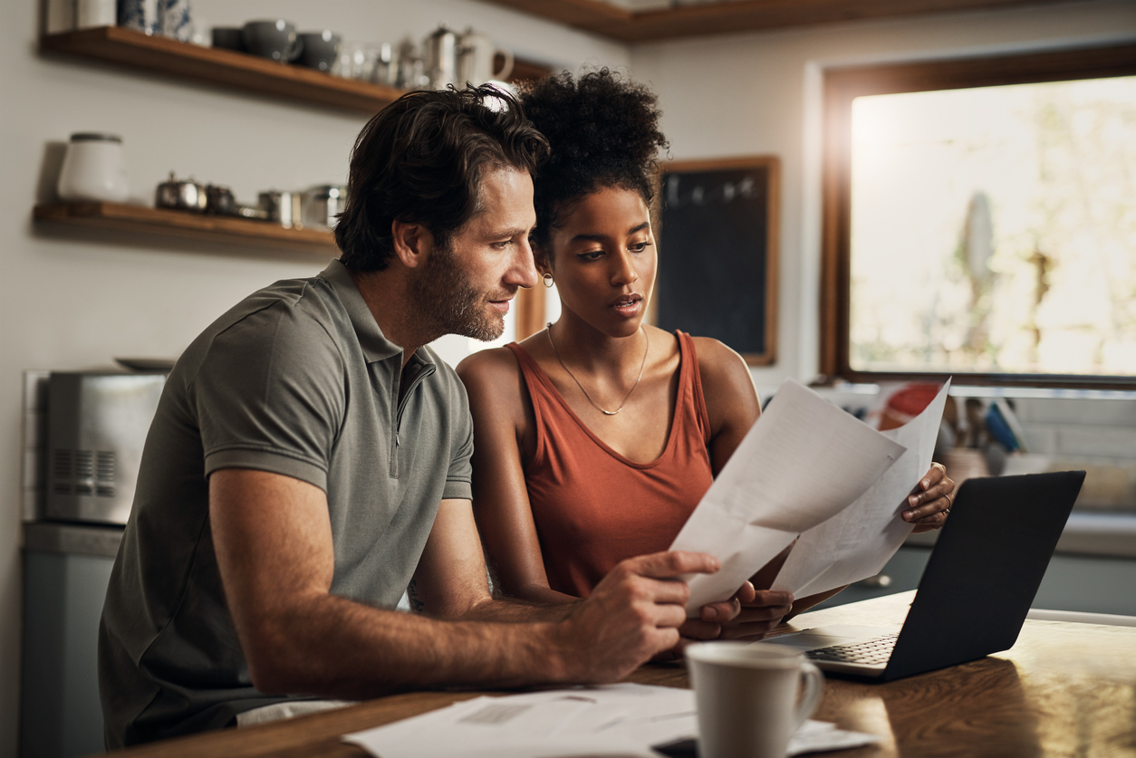 Couple sitting at table looking over a stack of bills. They have a neutral expression.