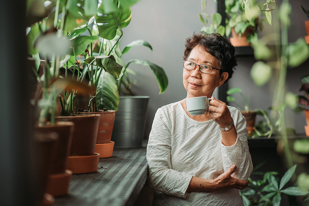 Older Asian woman enjoying a cup of coffee surrounded by plants