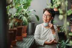 Older Asian woman enjoying a cup of coffee surrounded by plants